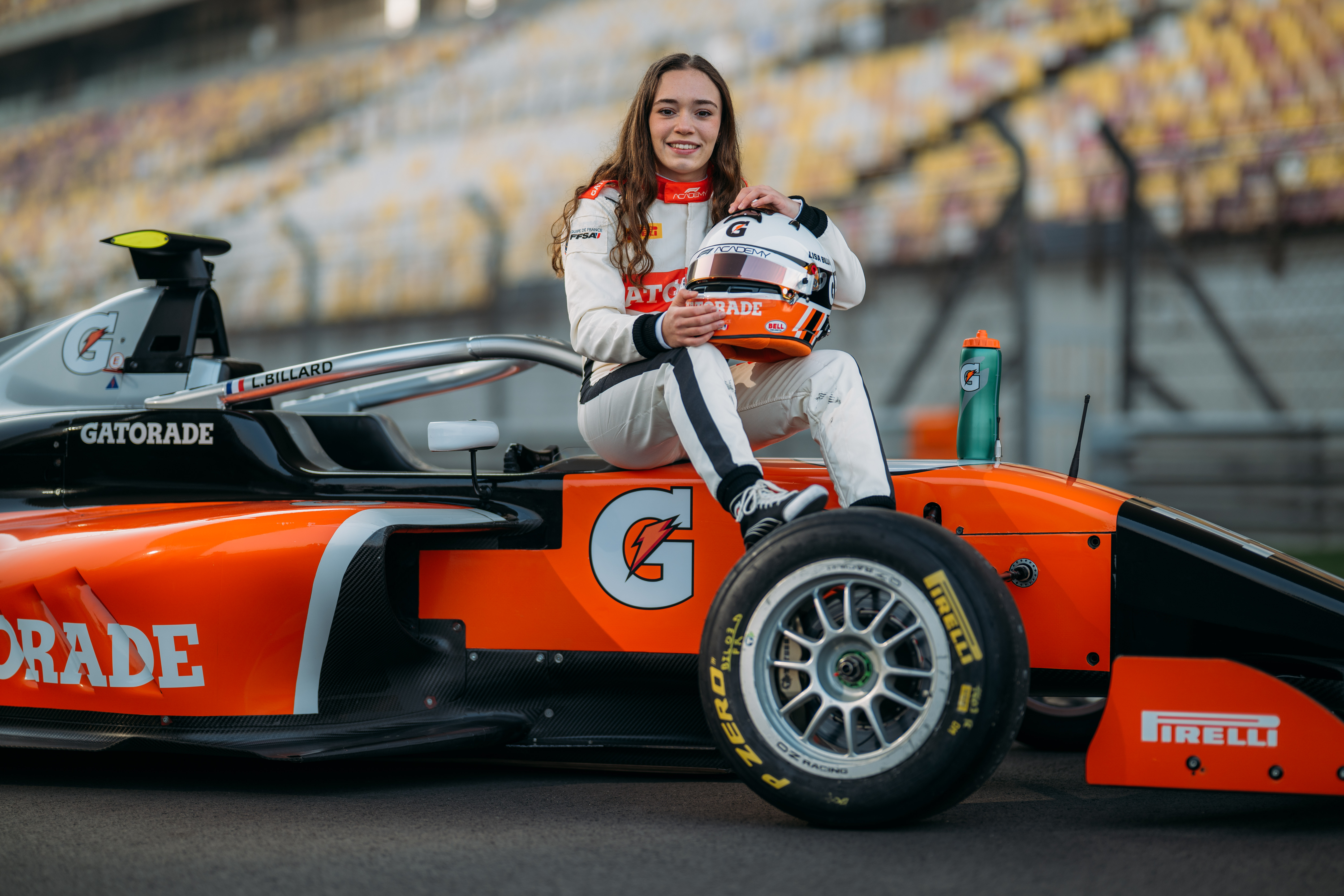 Pilote de course assis sur une monoplace orange aux couleurs de Gatorade, tenant un casque sur un circuit, avec des gradins de stade en arrière-plan.