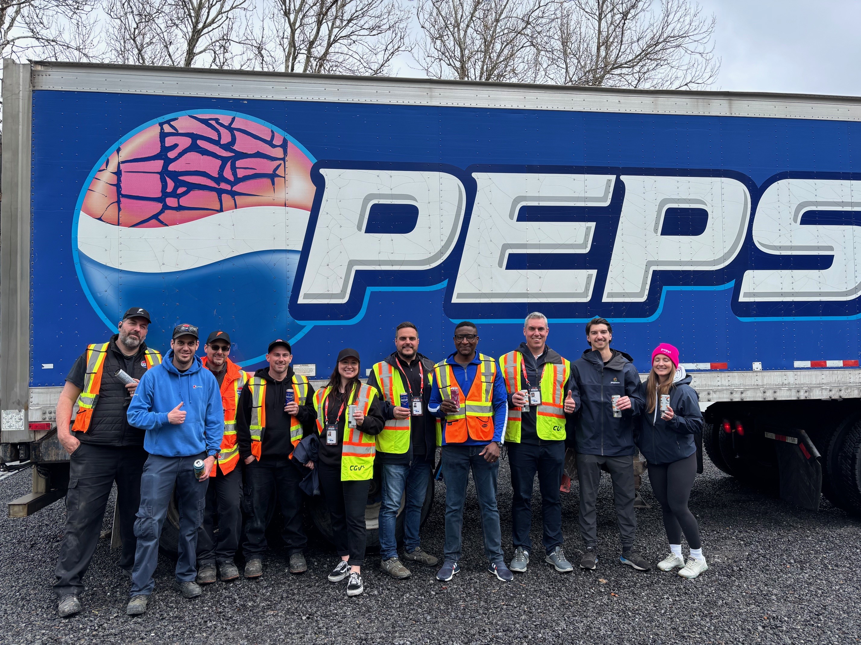 Membres de l’équipe PepsiCo portant des vestes de sécurité posent ensemble en tenant des boissons devant un grand camion de transport aux couleurs de Pepsi, à l’extérieur.